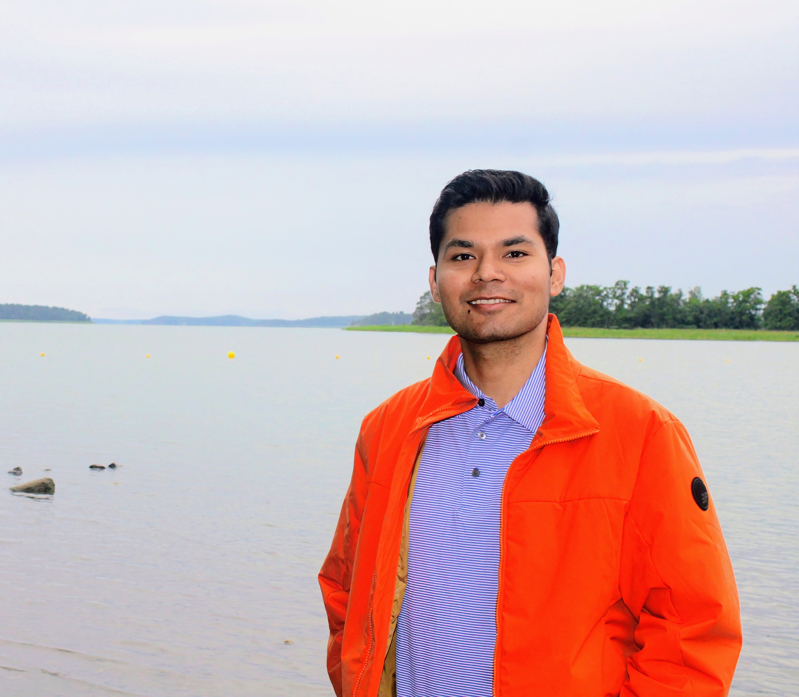 Shivam smiling in an orange jacket beside a calm lake, with a tree-lined shore in the background.