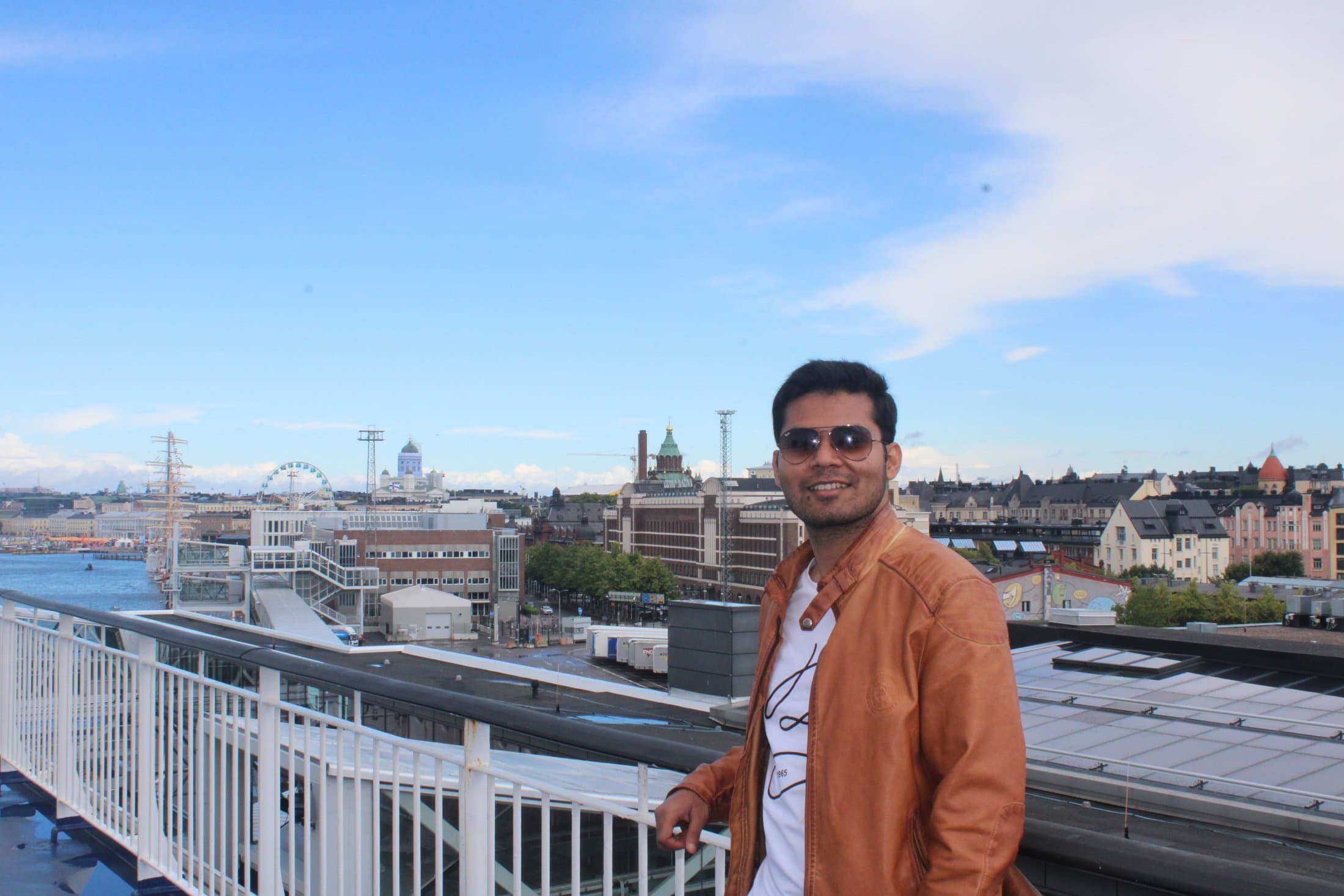 Shivam on an elevated walkway overlooking a Finnish harbor town with a Ferris wheel and waterfront buildings under a bright blue sky.