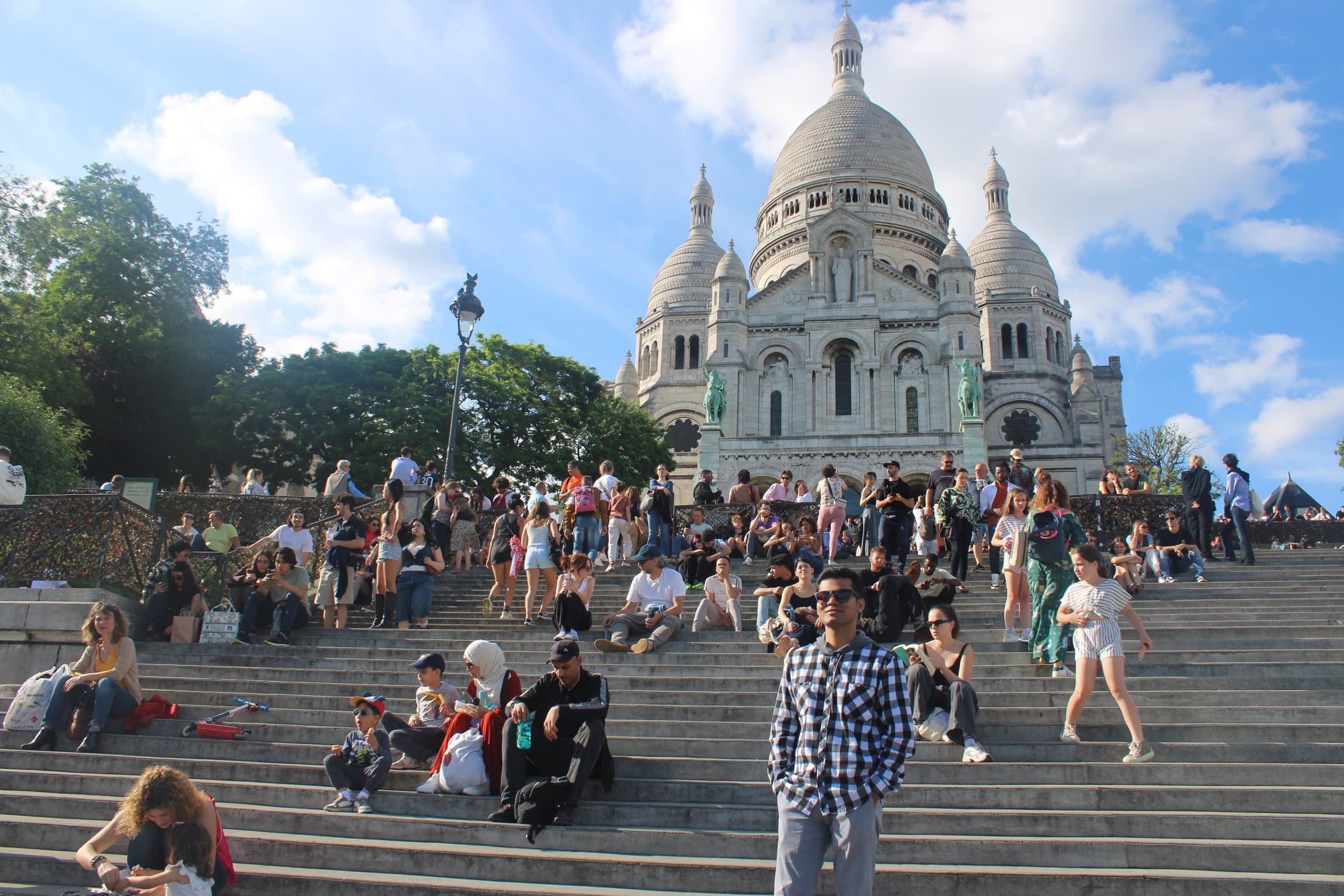 Shivam standing on the steps below the Sacré-Cœur Basilica in Montmartre, Paris, with the domes of the basilica rising behind a crowd on the steps.